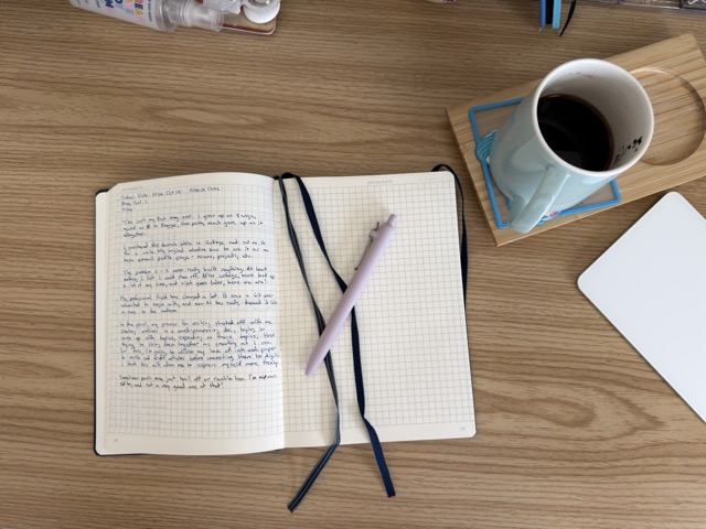 an open notebook sitting on my desk next to a cup of copy with a gel pen sitting on top. On the notebook's page is a written draft of this very blog post.